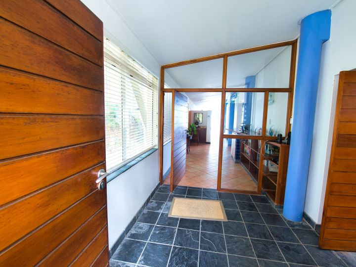 Large wooden door to the left of the image. Dark grey floor tiles. White walls. Blue pillar to the right side. Another open wooden door to the front and right.