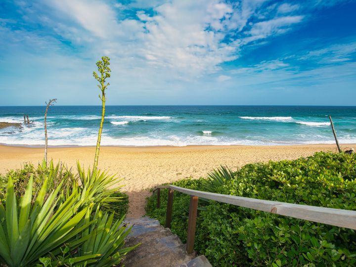 View of the ocean and beach front from the private access stairway. The sky is blue. It is partially cloudy, with white clouds. The ocean is a teal colour.