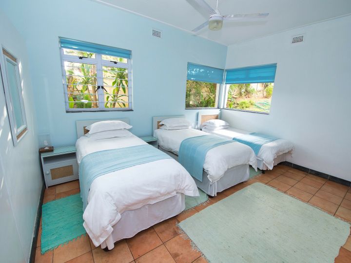 This is a room with one sky blue wall, and the remaining 3 white walls. There is a ceiling fan and three single beds. All three beds consisting of white linen and a turquoise throw-over