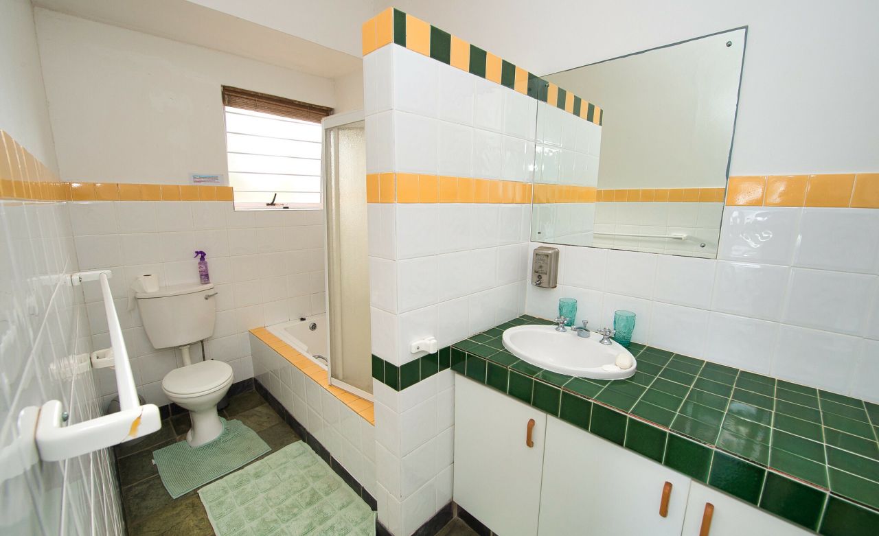 A bathroom with white tiled walls. The basin area is a dark green. There is a line of yellow tiles running horizontally through the centre of the walls. The mats on the floor are also green. A toilet is also visible.
